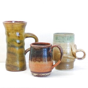May include: Three handcrafted ceramic mugs in earthy tones. The mugs vary in shape and size, with handles for easy gripping. The colors include shades of brown, green, and orange, with a rustic, textured finish. Perfect for coffee or tea.