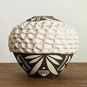 May include: A handcrafted ceramic vase with a unique design. The upper portion features a textured, wave-like pattern in a cream color. The lower section showcases a black and white geometric design. The vase sits on a wooden surface.