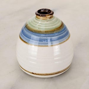 May include: A small, rounded ceramic vase with a narrow neck and a glossy finish. The body features horizontal bands of color, transitioning from a creamy white base to a vibrant blue middle section, topped with a pale green and brown rim.