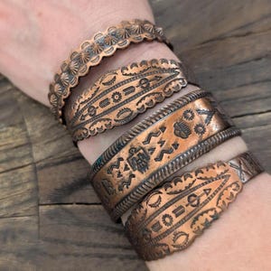 May include: A collection of copper-toned metal bracelets with intricate embossed designs. The bracelets feature scalloped edges and various patterns, including arrows, geometric shapes, and stylized bird motifs. The bracelets are stacked on a wrist, showcasing the different textures and styles.