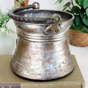 May include: A metallic bucket with a wide, flared rim and a tapered body featuring visible copper and silver-toned patina. It includes a dark, textured, arched metal handle attached to the sides with ornate, brass-colored metal brackets.