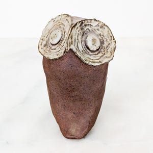 May include: A ceramic owl figurine featuring a dark brown, speckled body and two large, circular, off-white textured eyes with raised centers. The sculpture has a smooth, tapered form and a matte finish, showcasing a hand-crafted appearance.