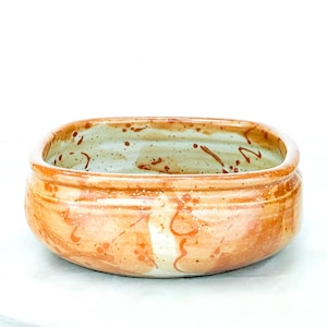 May include: A round ceramic bowl with a glossy finish, featuring a warm tan and light brown base color. The exterior and interior surfaces are decorated with scattered dark brown speckles and fluid, abstract brushstroke patterns in orange.