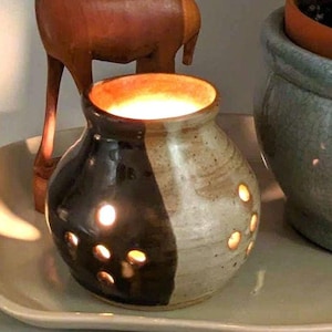May include: A ceramic candle holder with a two-tone design, black and beige. The candle holder has several small, round openings and is lit from within, creating a warm glow. The candle holder is on a white tray.