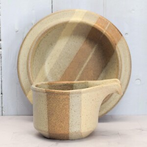 May include: A ceramic serving set featuring a plate and a gravy boat. The pieces have a speckled, beige finish with vertical stripes in shades of beige and brown. The plate is round, and the gravy boat has a handle.