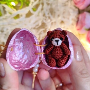 May include: A miniature dark brown crocheted teddy bear sits inside a hollowed-out walnut shell painted with a metallic pink interior. The bear features black bead eyes and a white stitched muzzle with a black nose. A small string of white pearls hangs.
