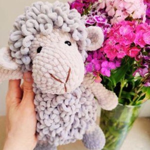 May include: A handmade, plush sheep toy in shades of gray and cream. The sheep has a fluffy, textured body and a stitched face. A bouquet of pink and purple flowers in a glass vase is in the background.