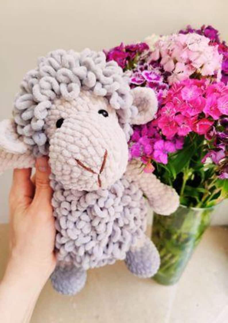 May include: A handmade, plush sheep toy in shades of gray and cream. The sheep has a fluffy, textured body and a stitched face. A bouquet of pink and purple flowers in a glass vase is in the background.