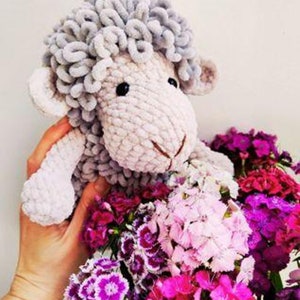 May include: A handmade, plush sheep toy with gray wool-like curls and a light pink body. The sheep is held over a bouquet of vibrant pink, magenta, and white flowers, creating a soft and colorful contrast.