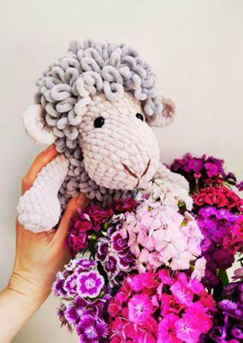 May include: A handmade, plush sheep toy with gray wool-like curls and a light pink body. The sheep is held over a bouquet of vibrant pink, magenta, and white flowers, creating a soft and colorful contrast.