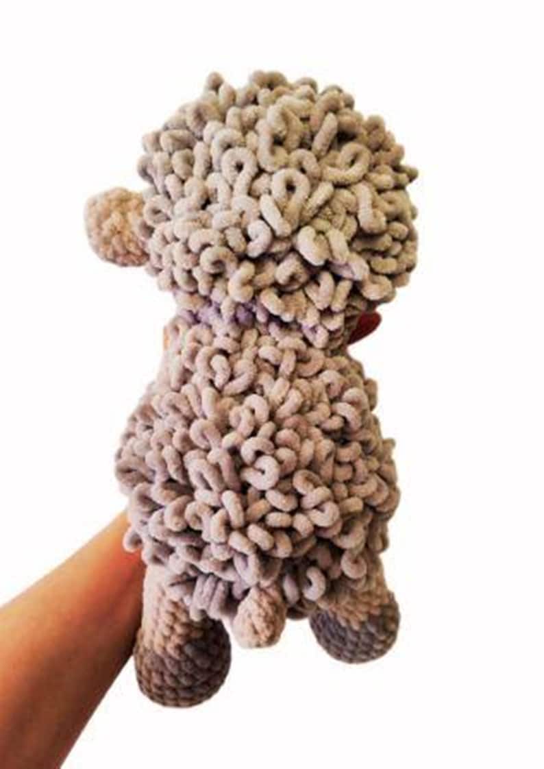 May include: A handmade, plush sheep toy in shades of gray. The sheep has a curly, textured body, small ears, and four stubby legs. The toy is held by a person's hand against a white background. The sheep is approximately 8 inches tall.