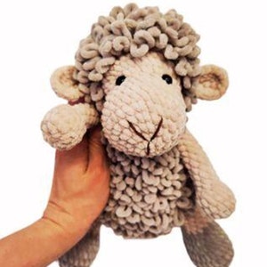 Puede incluir: Un peluche de oveja con un cuerpo beige claro y una cabeza texturizada y esponjosa. La oveja tiene pequeños ojos negros y una boca cosida. El juguete está sostenido por una mano.