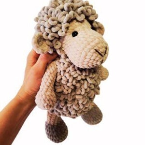 May include: A handmade plush sheep toy in shades of beige and gray. The sheep has a fluffy, curly head, small ears, and a stitched face. The body is made of a textured, looped yarn. The toy is being held by a person.