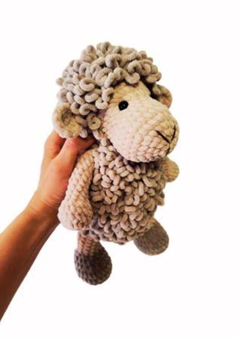 May include: A handmade plush sheep toy in shades of beige and gray. The sheep has a fluffy, curly head, small ears, and a stitched face. The body is made of a textured, looped yarn. The toy is being held by a person.