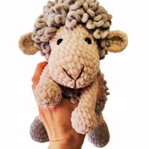 May include: A handmade plush sheep toy with a light gray, textured body and head. The sheep has small, rounded ears, black eyes, and a stitched smile. The toy is held up against a white background.