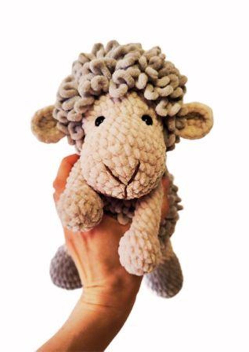 May include: A handmade plush sheep toy with a light gray, textured body and head. The sheep has small, rounded ears, black eyes, and a stitched smile. The toy is held up against a white background.