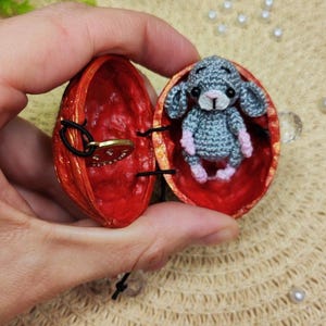 May include: A small gray crocheted mouse figure with pink accents sits inside a hollowed-out walnut shell painted bright red on the interior. A small gold-toned circular charm with a smiley face design is attached to the other side of the shell.