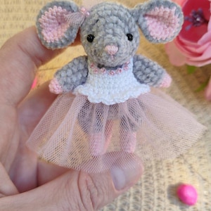 May include: A handcrafted grey crochet mouse wearing a white top, pink tutu, and pink ballet slippers. The mouse has pink-lined ears and a pink nose. A small pink bow adorns the top of its head. The mouse is a whimsical, handcrafted item.