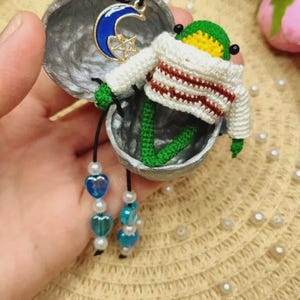 May include: A small green crocheted frog figurine wearing a white sweater with brown stripes, nestled inside a silver-painted walnut shell. The frog has dangling legs adorned with white pearls and blue heart-shaped beads, plus a blue moon charm.