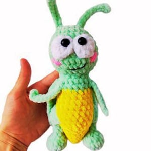 May include: A crochet plush toy shaped like a green insect with two antennae, large white eyes with black pupils, and pink cheeks. The creature has a textured light green body and a bright yellow belly, crafted from soft yarn with visible stitches.