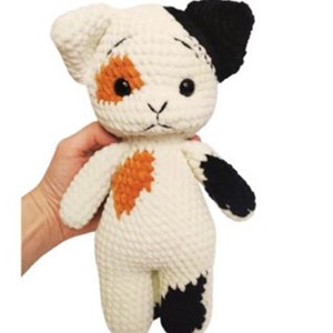 Puede incluir: Muñeca de gato de ganchillo hecha a mano. La muñeca tiene un cuerpo blanco con marcas negras y naranjas. Tiene ojos negros de botón y está siendo sostenida. La muñeca de gato es un juguete blando.