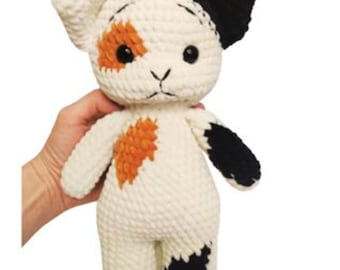 May include: A handmade crochet cat doll with a white body, black ear, and orange and black markings. The doll has black button eyes and is being held by a hand. The cat doll is a soft toy.
