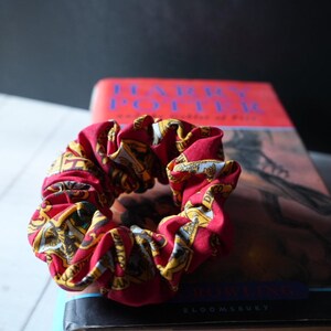 Peut inclure: Un chouchou en tissu rouge orné d'un motif répétitif de blasons et de textes dorés et noirs. Le chouchou est posé sur un livre Harry Potter à couverture rouge, affichant le titre en lettres bleues ainsi que le nom de l'auteur.