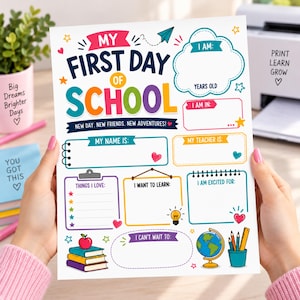 以下が含まれることがあります： My First Day of Schoolというテキストとカラフルなグラフィックが描かれた白い紙のサイン。名前、年齢、先生、興味、目標を記入するセクションがあり、本、リンゴ、地球儀、鉛筆のイラストで装飾されています。