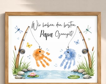 May include: A framed print featuring two handprint fish in orange and blue, each caught on a fishing rod. The text reads Wir haben den besten Papa Geangelt! The scene includes a pond with water, rocks, a pink water lily, dragonflies, and butterflies.