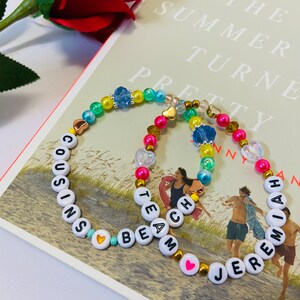 Puede incluir: Dos pulseras de cuentas coloridas con cuentas de letras blancas que deletrean los nombres "Cousins Beam" y "Jeremiah". Las pulseras est&aacute;n sobre un libro con el t&iacute;tulo "The Summer I Turned Pretty" visible.