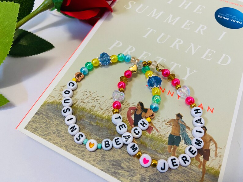 Puede incluir: Dos pulseras de cuentas coloridas con cuentas de letras blancas que deletrean los nombres "Cousins Beam" y "Jeremiah". Las pulseras est&aacute;n sobre un libro con el t&iacute;tulo "The Summer I Turned Pretty" visible.
