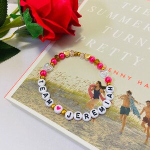 Puede incluir: Un brazalete de cuentas rosadas y doradas con cuentas de letras blancas que deletrean "Team Jeremiah" con un dije de coraz&oacute;n rosa. El brazalete est&aacute; sobre un libro titulado "The Summer I Turned Pretty" de Jenny Han.