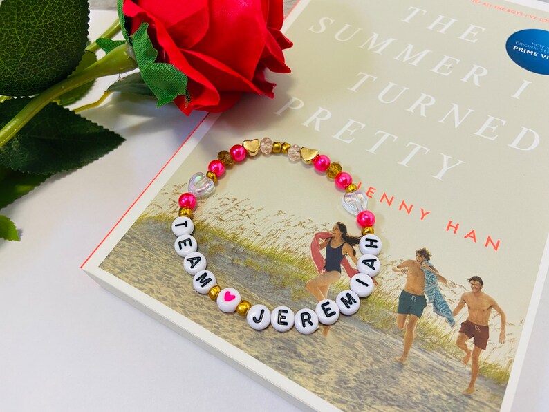 Puede incluir: Un brazalete de cuentas rosadas y doradas con cuentas de letras blancas que deletrean "Team Jeremiah" con un dije de coraz&oacute;n rosa. El brazalete est&aacute; sobre un libro titulado "The Summer I Turned Pretty" de Jenny Han.
