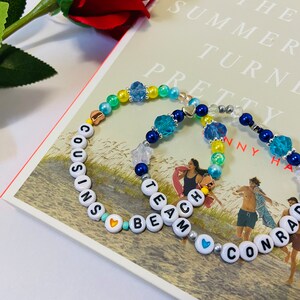 Puede incluir: Dos pulseras de cuentas con las palabras "COUSINS BEACH" y "TEAM CONRAD" escritas en cuentas blancas. Las pulseras est&aacute;n hechas con cuentas azules, amarillas y verdes y tienen peque&ntilde;os corazones y estrellas plateados.