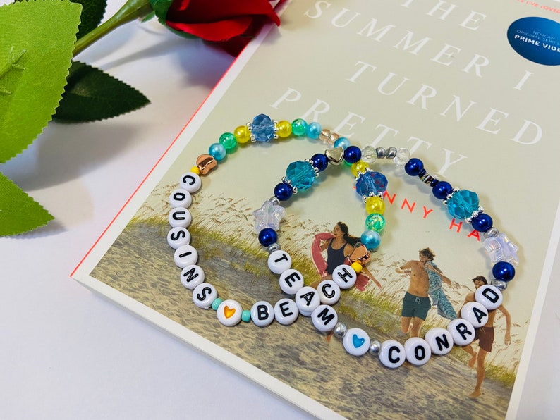 Puede incluir: Dos pulseras de cuentas con las palabras "COUSINS BEACH" y "TEAM CONRAD" escritas en cuentas blancas. Las pulseras est&aacute;n hechas con cuentas azules, amarillas y verdes y tienen peque&ntilde;os corazones y estrellas plateados.