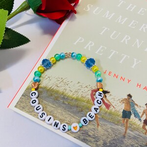 Puede incluir: Un brazalete de cuentas de colores con las palabras "COUSINS" y "BEACH" escritas en letras blancas. El brazalete tiene cuentas azules, verdes y amarillas, as&iacute; como peque&ntilde;os dijes de coraz&oacute;n dorados.