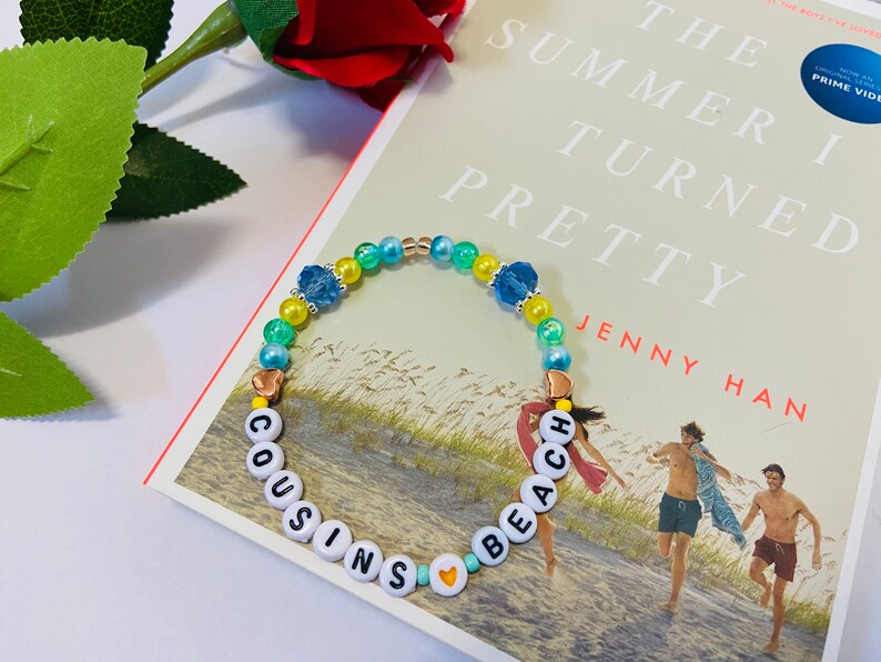 Puede incluir: Un brazalete de cuentas de colores con las palabras "COUSINS" y "BEACH" escritas en letras blancas. El brazalete tiene cuentas azules, verdes y amarillas, as&iacute; como peque&ntilde;os dijes de coraz&oacute;n dorados.