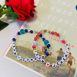 Puede incluir: Dos pulseras de cuentas con cuentas de letras blancas que deletrean "Team Conrad" y "Team Jeremiah" con cuentas azules, rosadas y doradas. Las pulseras est&aacute;n sobre un libro titulado "The Summer I Turned Pretty" de Jenny Han.