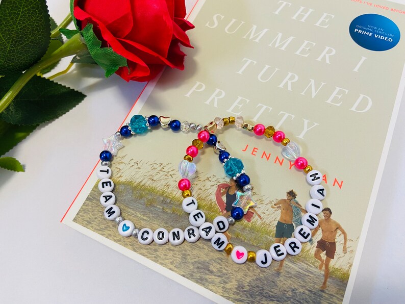 Puede incluir: Dos pulseras de cuentas con cuentas de letras blancas que deletrean "Team Conrad" y "Team Jeremiah" con cuentas azules, rosadas y doradas. Las pulseras est&aacute;n sobre un libro titulado "The Summer I Turned Pretty" de Jenny Han.