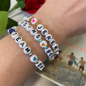 Puede incluir: Tres pulseras de cuentas blancas con abalorios en forma de coraz&oacute;n de colores. Las pulseras deletrean los nombres "TEAM", "JEREMIAH" y "AUSTIN".