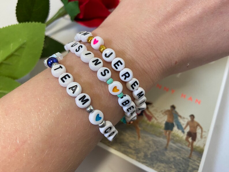 Puede incluir: Tres pulseras de cuentas blancas con abalorios en forma de coraz&oacute;n de colores. Las pulseras deletrean los nombres "TEAM", "JEREMIAH" y "AUSTIN".