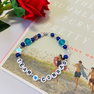 Puede incluir: Un brazalete de cuentas azules y plateadas con cuentas de letras blancas que deletrean "Team Conrad" y abalorios de estrellas y corazones azules y plateados. El brazalete est&aacute; encima de un libro titulado "The Summer I Turned Pretty" de Jenny Han.