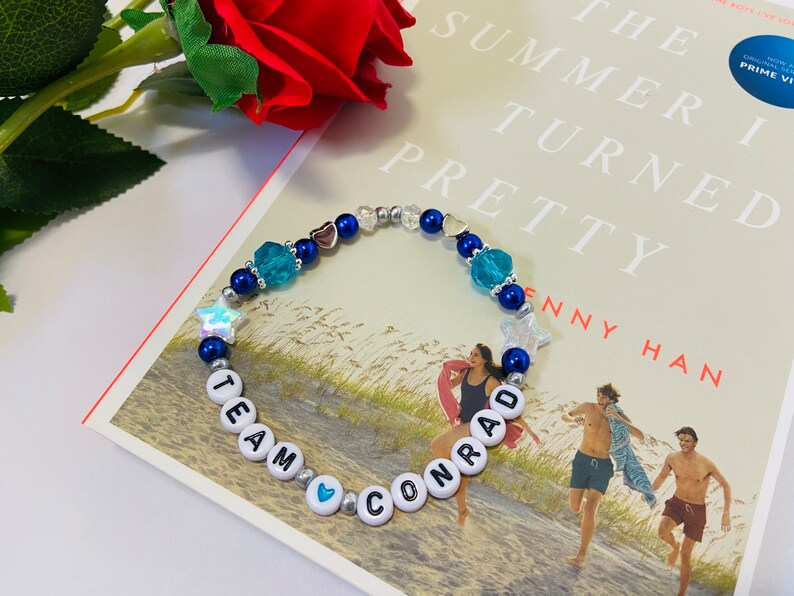 Puede incluir: Un brazalete de cuentas azules y plateadas con cuentas de letras blancas que deletrean "Team Conrad" y abalorios de estrellas y corazones azules y plateados. El brazalete est&aacute; encima de un libro titulado "The Summer I Turned Pretty" de Jenny Han.