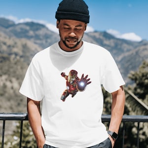 K&ouml;nnte beinhalten: Ein wei&szlig;es T-Shirt mit einem roten und goldenen Iron Man-Motiv auf der Vorderseite. Das Motiv zeigt Iron Man im Flug mit ausgestreckter Hand, der einen Repulsorstrahl abfeuert.