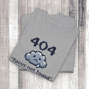 May include: Two light gray t-shirts are stacked on a wooden surface. The top shirt features a pixelated design with the number "404" above a sad cloud graphic. Below the cloud, the text reads "focus not found".