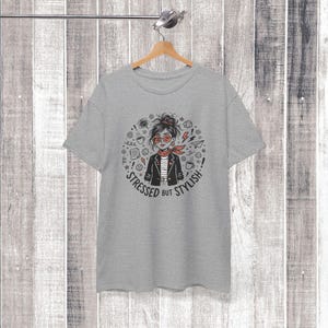 May include: A grey t-shirt with a cartoon illustration of a woman wearing red glasses, a red scarf, and a black jacket. The text "Stressed but Stylish" is printed around the illustration. The t-shirt is hanging on a wooden hanger.