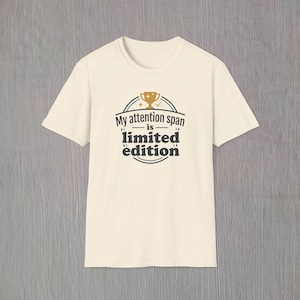Może przedstawiać: Kremowa koszulka z napisem "My attention span is limited edition" w kolorze czarnym i złotą grafiką trofeum. Koszulka ma okrągły dekolt i krótkie rękawy, odpowiednia do noszenia na co dzień.