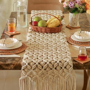 May include: A cream macrame table runner with a geometric design and fringed edge. The table is set with a fruit basket, a glass jug, and flowers. Coasters, plates, and glasses complete the dining scene.