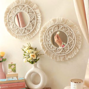 May include: Two round, cream-colored macrame mirrors with fringe hang on a white wall. A white vase with a circular opening holds flowers. A stack of books and a candle sit on a wooden shelf below the mirrors. The mirrors reflect the room.