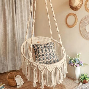 Puede incluir: Una silla colgante de macramé color crema con un cojín estampado. La silla tiene un diseño circular con flecos. El fondo incluye una alfombra tejida, una cesta y adornos de pared, creando una estética bohemia.